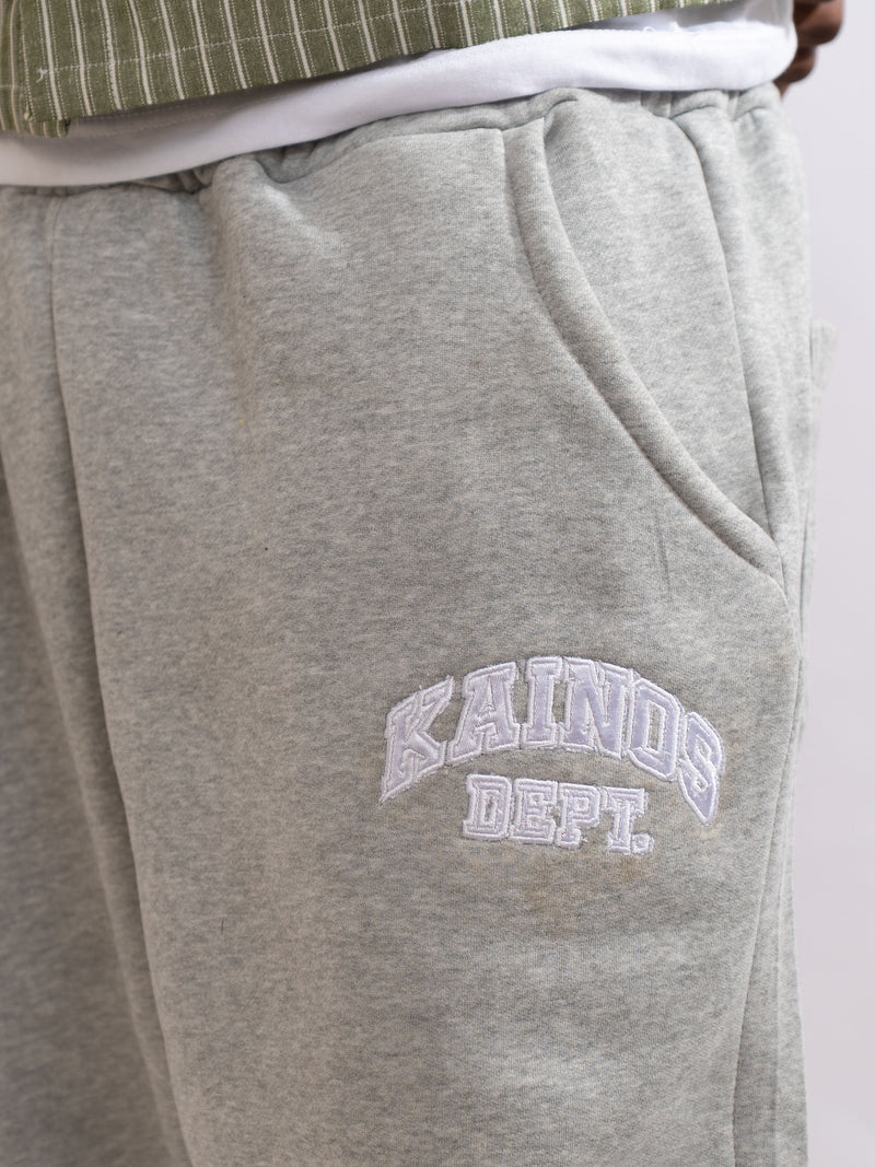 Close-up of the grey sweatpants worn with a striped shirt, showing the KAINOS DEPT. varsity print on the thigh and a side pocket