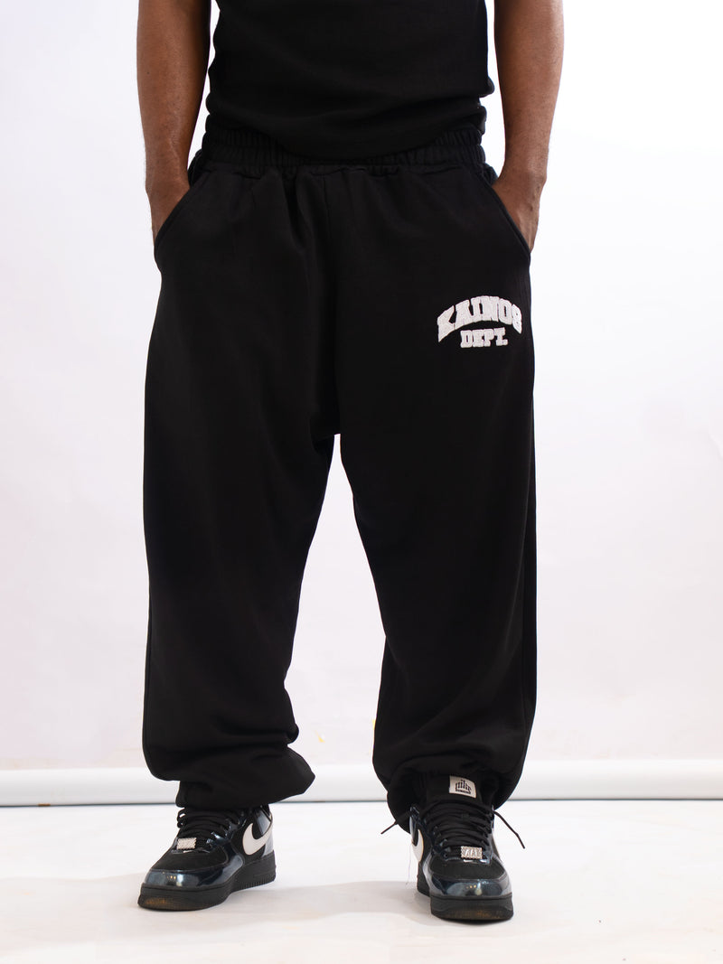 Model wearing the black sweatpants with hands in the side pockets, showing the KAINOS DEPT. print on the thigh